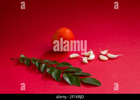 Spicchi d'aglio con foglie di curry e pomodoro, arrangiati splendidamente su uno sfondo rosso testurizzato, isolati. Foto Stock