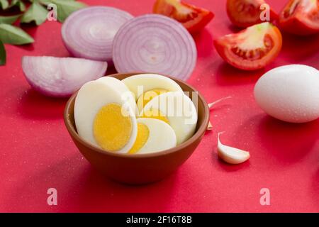 Ingredienti crudi per la cottura del masala d'uovo, cipolla, pomodoro, foglie di curry, aglio e uovo bollito disposti su uno sfondo rosso Foto Stock