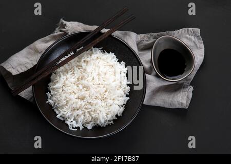 Riso bianco bollito in stile asiatico in ciotola nera con chopsticks e salsa di soia sul lato. Sfondo scuro. Vista dall'alto Foto Stock