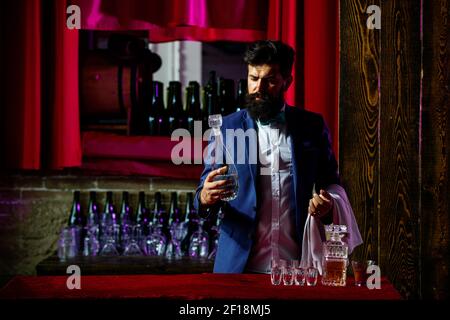 Bel barman con barba lunga e baffi con cocktail alcolico sul viso in pelle scamosciata vintage su sfondo viola rosso bar. Foto Stock