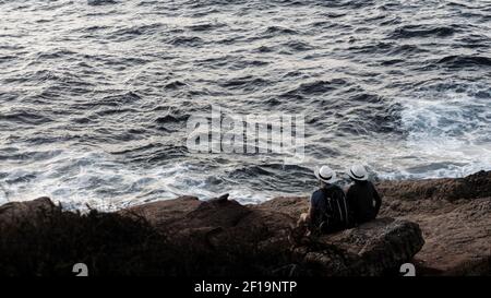 Giovane coppia romantica in bianco cappelli identici con uno zaino stanno guardando il tramonto mentre si siede sulla costa rocciosa. I raggi del sole che tramonta Foto Stock