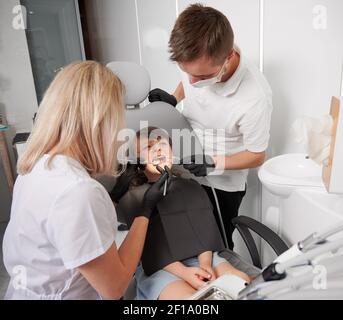 Bambino seduto in sedia dentale mentre due dentisti che controllano i denti del bambino. Dentista che esamina i denti della ragazza mentre l'assistente maschio tiene l'iniettore della saliva. Concetto di odontoiatria pediatrica e cura dentale. Foto Stock