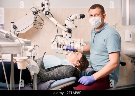 Ritratto di dentista maschile che tratta i denti della donna in clinica dentale. Stomatologo che guarda la macchina fotografica e tiene gli strumenti dentali mentre si siede accanto al paziente. Concetto di odontoiatria e cura dentale. Foto Stock