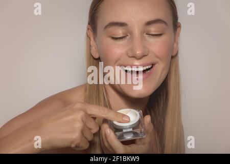 Ritratto di una donna sorridente felice usa crema facciale. Faccia isolata su sfondo beige. Prodotto anti-invecchiamento o antirughe. Bellezza e cura della pelle Foto Stock