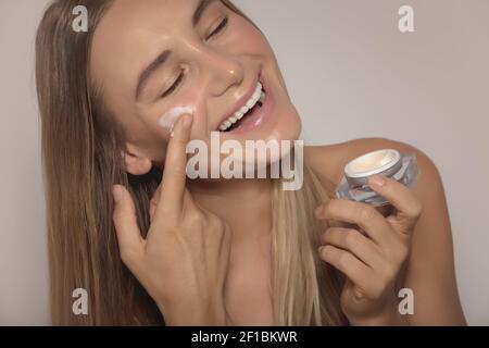 Ritratto di un felice modello sorridente applicando crema facciale. Isolato su sfondo beige. Cosmetici anti-invecchiamento o anti-rughe. Pelle perfetta e liscia Foto Stock