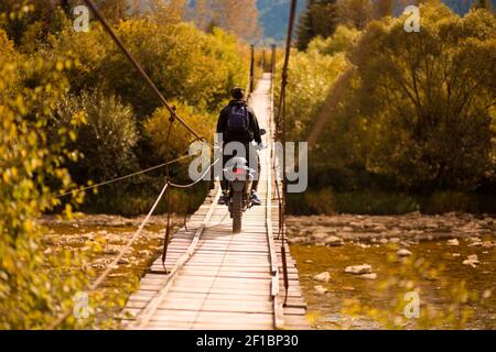 Uomo irriconoscibile che cammina con moto da fondo sul ponte sospeso oltre le montagne fiume in estate Foto Stock