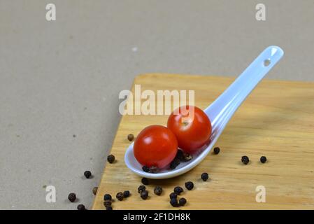 Due piccoli pomodori rossi su un cucchiaio di porcellana asiatica. Sfondo marrone chiaro con tavola di legno e palle di pepe nero. Decorazione di vegetabl fresco Foto Stock