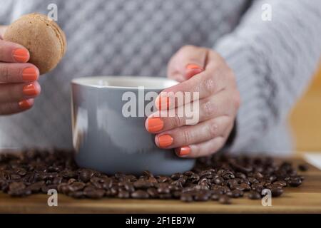 Mano femminile che tiene una tazza di caffè e un macaroon. Chicchi di caffè su una tavola di legno. Dolce dessert. Foto Stock