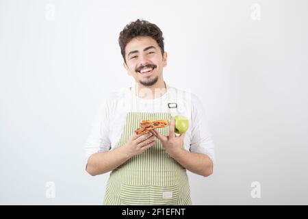 Ritratto del cuoco maschio sorridente che tiene mela e pizza sopra sfondo bianco Foto Stock