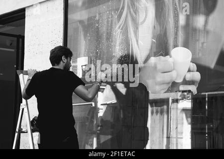 PARIGI, FRANCIA - 1 SETTEMBRE 2018: Giovane uomo che rimuove il vecchio poster pubblicitario con la donna che tiene una tazza di caffè dalla vetrina del negozio. Foto in bianco e nero Foto Stock