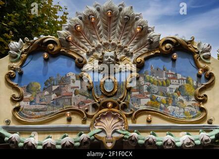 Dipinti di tipiche città provenzali e villaggi riempiono pannelli sulle tavole ornamentali arrotondate fissate in cima a una tradizionale giostra d'epoca che offre giostre ai giovani dalla Fontaine de la Rotonde in Place Général de Gaulle a Aix-en-Provence, Bouches-du-Rhône, Provence-Alpes-Côte d'Azur, Francia. Molti caroselli o giostre ben restaurati e mantenuti nel fairground costruito nell'era della Belle Epoque alla fine del 1800 e all'inizio del 1900 sono ancora in funzione in tutta la Francia moderna. Foto Stock