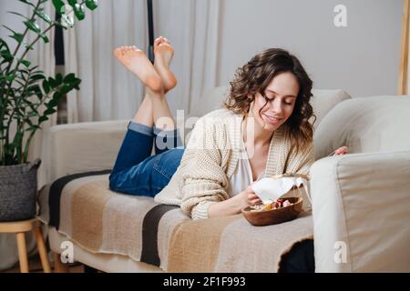 Felice giovane donna cucita nel comfort del suo appartamento. È sdraiata su un divano, ricamando un'immagine colorata su un loop. Foto Stock