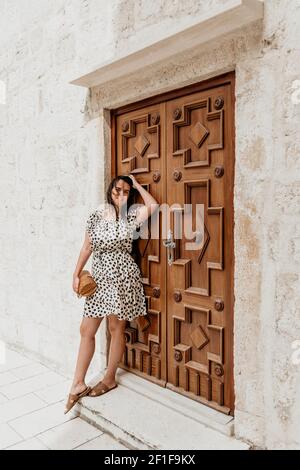 Una donna caucasica sorridente in un abito fiorito punteggiato di polka in posa contro una vecchia porta decorativa Foto Stock