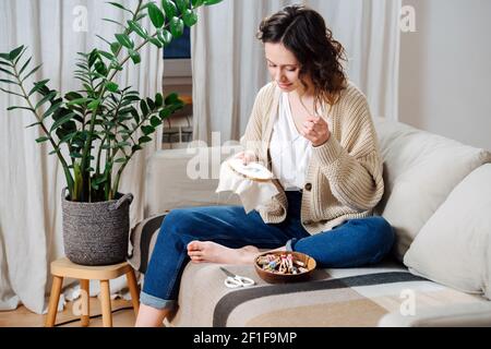 Felice giovane donna cucita nel comfort del suo appartamento. È seduta su un divano, ricamo un'immagine colorata su un loop. Foto Stock