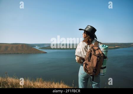 Bella giovane donna con zaino in piedi in cima alla collina e godere Dniester canyon. Donna turistica in cappello cowboy trascorrere il tempo libero attivamente all'aperto. Foto Stock