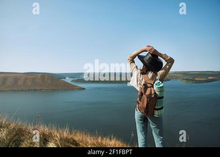 Giovane donna in abito casual e cappello cowboy godendo di una bella vista del fiume Dniester da alta collina. Donna piacevole con zaino trekking parco nazionale Podillya Tovtry. Foto Stock