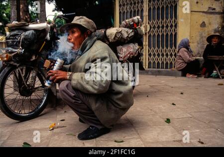 Un ciclista itinerante fumo oppio in un angolo di strada, Hanoi, Vietnam, giugno 1980 Foto Stock