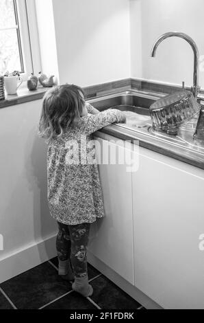 Ragazza giovane di 4 anni che si allunga fino a. lavatele le mani nel lavandino della cucina Foto Stock