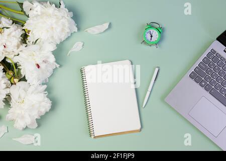 apri il notebook per scrivere piani, per fare lista o sogni con peonie fresche, computer portatile, sveglia, su sfondo verde chiaro Foto Stock