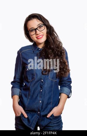 Ritratto di una giovane donna sicura con lunghi capelli ondulati e occhiali in camicia in denim sorridente con le mani nelle tasche jean. Isolato su bianco Foto Stock