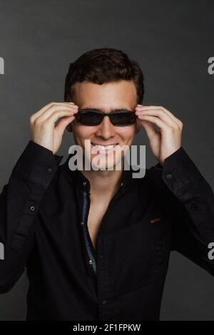un giovane uomo dai capelli scuri si siede su uno sfondo nero in una camicia nera, indossando occhiali da sole Foto Stock