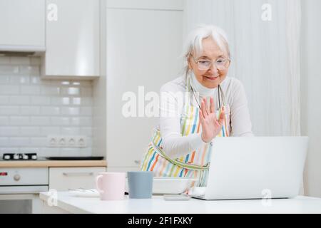 Una donna anziana ondeggia mentre, salutando e chattando via videochiamata in un computer portatile. Si trova in cucina in un grembiule Foto Stock