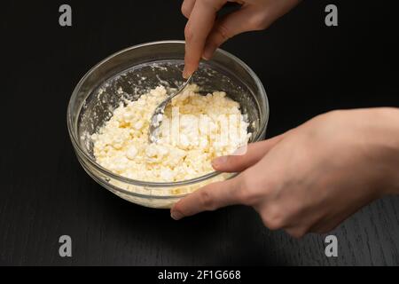 Le mani delle donne mescolano un cucchiaio in una ciotola di vetro di formaggio di cottage granuloso, uovo, farina su un primo piano del tavolo nero Foto Stock