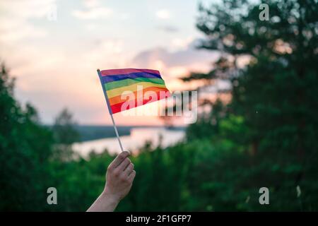 Una mano ondeggia un colorato gay orgoglio LGBT arcobaleno bandiera al tramonto su un paesaggio naturale in estate. Foto Stock