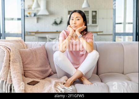 Giovane adulto asiatico misto-razza casalinga femminile con lunghi capelli neri seduti sul divano, sentendosi annoiati e demotivati, non vuole fare compiti, sconvolto con il messaggio dal telefono, infelice Foto Stock