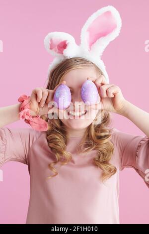 bambino elegante sorridente con lunghi capelli biondi ondulati con orecchie conigliate e uovo di pasqua su sfondo rosa. Foto Stock