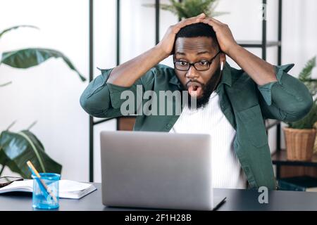 Sorpreso scioccato afroamericano, si siede alla sua scrivania, tenendo la testa con le mani, guardando un laptop, ha ricevuto una notizia o un messaggio inaspettato, ha visto una dichiarazione finanziaria, scioccato espressione facciale Foto Stock