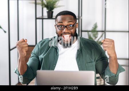 Esultante freelancer africano-americano vestito con abiti eleganti, seduto alla sua scrivania, gioendo nel successo, vittoria o buon profitto, guardando la macchina fotografica e gesturando con le mani Foto Stock