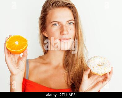 giovane donna bionda che sceglie tra ciambella e frutta arancio isolato su bianco, concetto di dieta Foto Stock