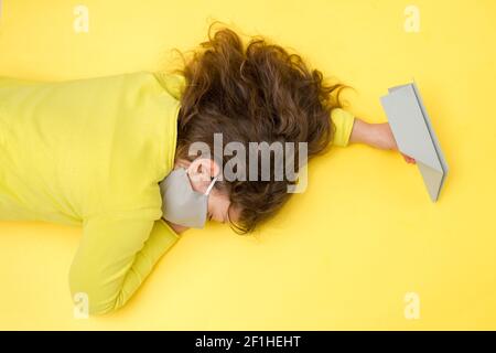 Addormentando la bambina in una maschera grigia protettiva in mano con un aeroplano di carta grigio su sfondo giallo. Concetto creativo di viaggio, vacanza durante Foto Stock