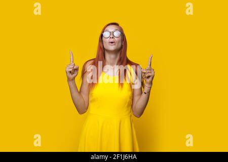 Donna zenzero sorpresa sta puntando su con entrambe le dita che posano su una parete di studio in un vestito giallo Foto Stock