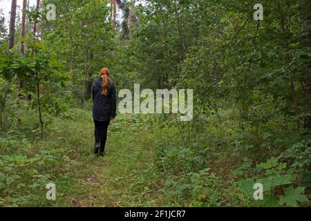 Una ragazza decapitata in una giacca nera e leggings è camminando lungo un sentiero sull'erba verde in un foresta di compensazione tra alberi alti con foglie succose Foto Stock