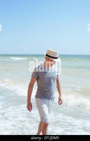 Un uomo in una camicia grigia, pantaloncini bianchi e cappello di paglia che cammina sulla spiaggia Foto Stock