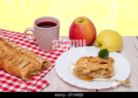 Apple roll servito. Un rotolo di strudel di mele fresco cotto in casa con una tazza di tè, una fetta appetitosa di torta di mele sul piatto e mele fresche su un Foto Stock