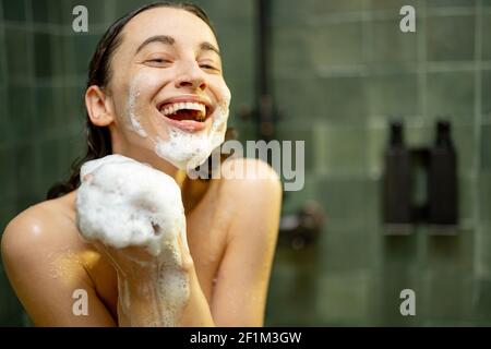 Donna sorridente che prende la doccia e che sorvola il corpo con gel doccia in bagno verde. Foto di routine quotidiana. Guardando la fotocamera. Foto Stock