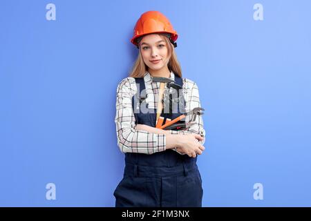 Donna sicura che indossa un casco arancione e una tuta blu, una chiave di fissaggio, un martello e altri strumenti in mani isolate su sfondo blu dello studio, Foto Stock