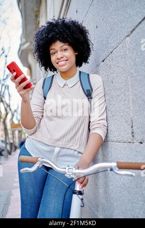 Donna afroamericana sorridente con uno smartphone in mano. Lei fa un giro in bicicletta attraverso la città. Foto di alta qualità Foto Stock