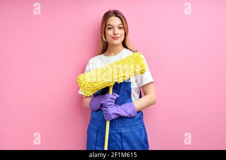 La cameriera sta per pulire il pavimento con il mop, stare in piedi in guanti di gomma che si pongono alla telecamera isolata su sfondo rosa, ritratto. Di donna in insenatura blu Foto Stock