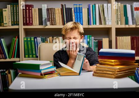ragazzino esausto che urla mentre studia, fa i compiti, seduto a tavola circondato da libri, in biblioteca Foto Stock