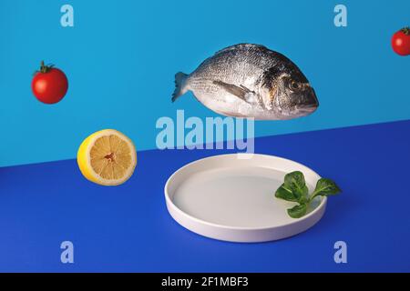 Pesce dorado antigravitazionale, foglie verdi e pomodori ciliegini volano su un piatto su uno sfondo blu luminoso Foto Stock