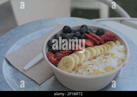 Un colpo di fuoco selettivo di una ciotola di Acai riempita con mirtilli, fragole e banane in una ciotola bianca di ceramica su un tavolo blu Foto Stock