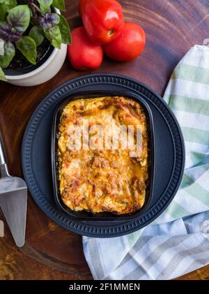 Deliziosa lasagna bolognese su piastra nera in ceramica, con posate, pomodori, vaso con basilico e panno a righe su base di legno. Vista dall'alto. Foto Stock