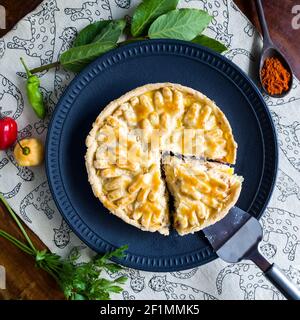 gustosa torta di pollo su piatto nero con peperoni gialli e rossi, foglie verdi, cucchiaio di legno, prezzemolo e paprika su sfondo di legno. Vista dall'alto. Foto Stock