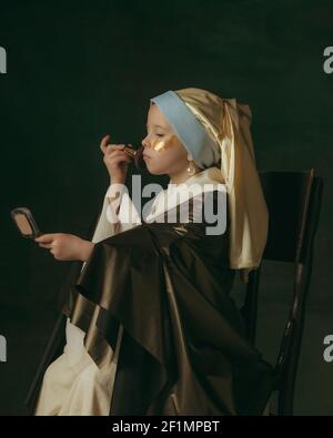 Usando le macchie dorate dell'occhio, polvere del viso. Bambina medievale come signora con un orecchino perla su sfondo scuro studio. Concetto di confronto di epoche, infanzia, antica. Elegante, creativa, visione artistica. Foto Stock