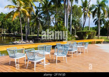 Sedie e tavoli bianchi e blu vuoti si trovano su una terrazza con pavimento in legno in una giornata di sole, palme da cocco sono sullo sfondo Foto Stock