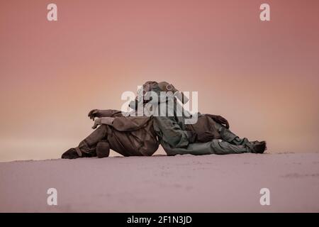mondo post-apocalittico dopo una guerra nucleare. Due uomini in maschere a gas e tute protettive si siedono sulla sabbia con le spalle l'una all'altra. Foto Stock